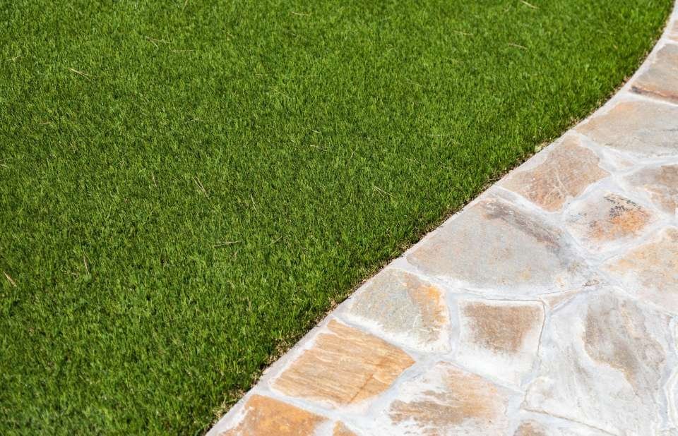 A close-up view of a neatly trimmed grassy area adjacent to a stone pathway. The image showcases the clear border between Scottsdale Artificial Turf and the light-colored, irregularly shaped paving stones, creating a pristine outdoor aesthetic.