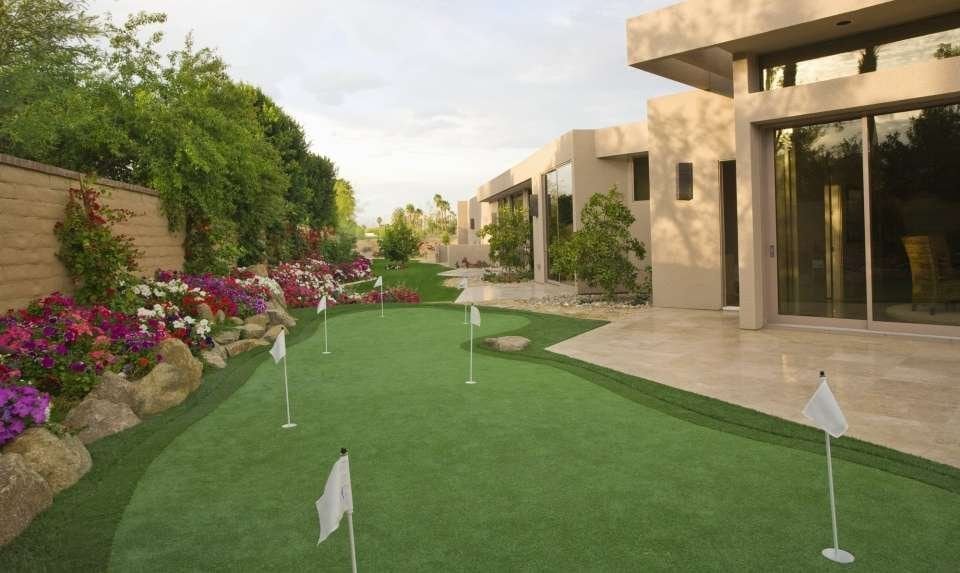 A backyard putting green with several small white flags, bordered by a vibrant garden of blooming flowers and shrubs. A modern house with large windows and a tiled patio is on the right, while a stone wall is on the left under a partly cloudy Paradise Valley sky.