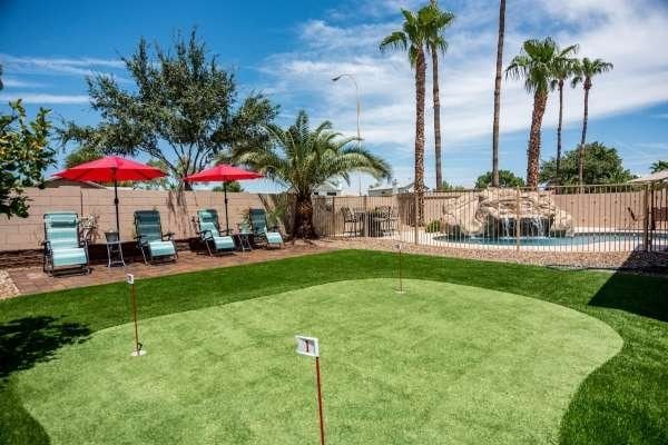 A backyard in Scottsdale, AZ, boasts a putting green installed by professional golf green installers. Sun loungers with red umbrellas rest beneath tall palm trees and lush plants. A stone waterfall feature stands near a fenced-in pool area under a clear blue sky, promising a perfect sunny day.