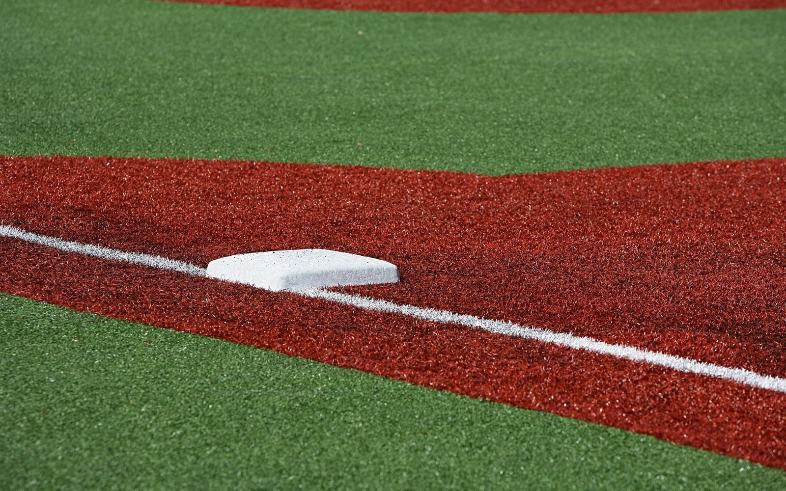 Close-up of a baseball field's base with synthetic grass. The base is white and positioned on a section of red artificial turf, with a white foul line running alongside it. Green turf surrounds the base and red section, creating a distinct contrast.