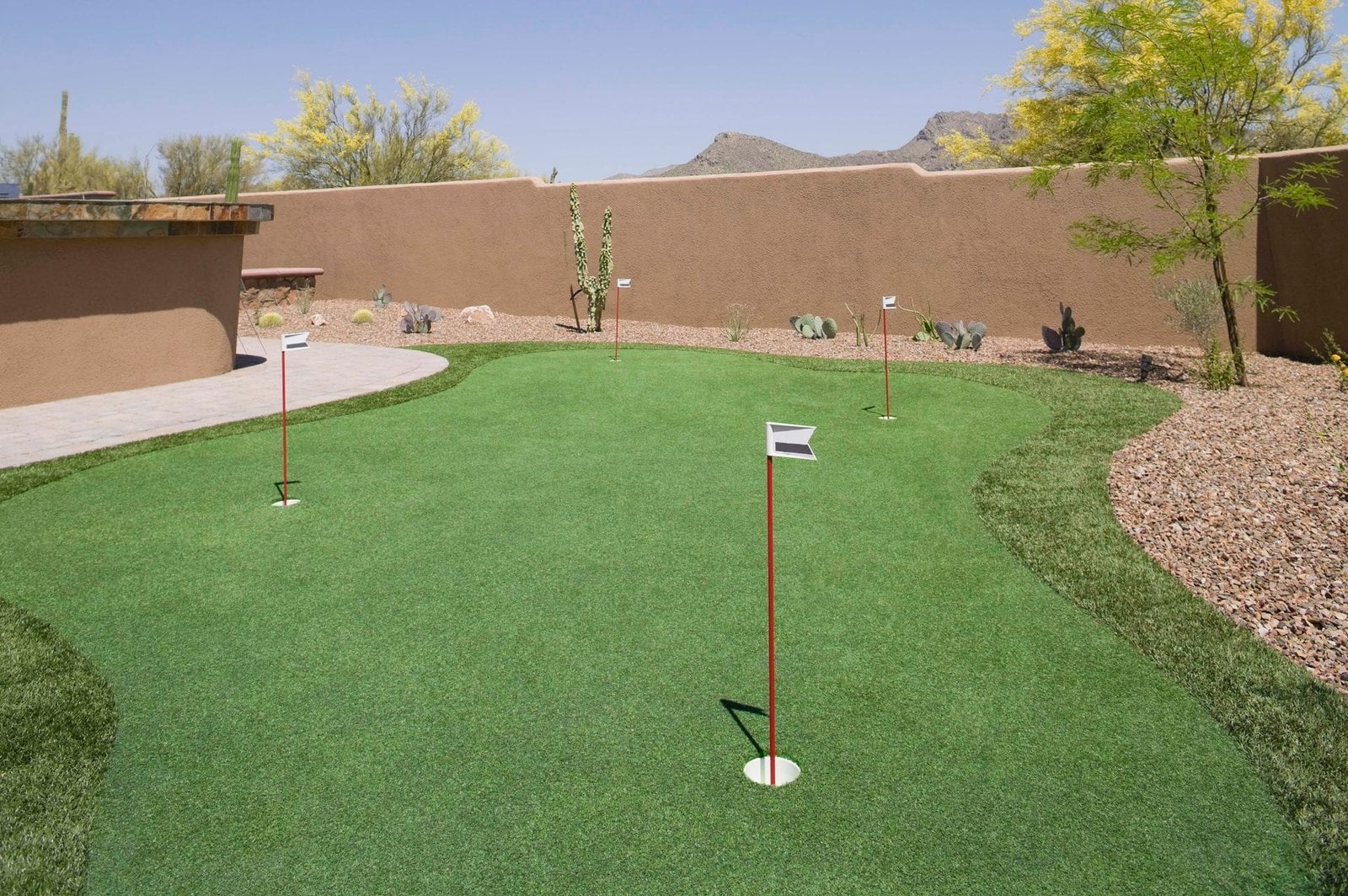 A well-maintained putting green with five marked holes, situated outdoors on artificial turf. The area is surrounded by a low stone wall and landscaped with gravel and desert plants. Trees and mountains are visible in the background under a clear sky. Licensed installers offer free consults for setup.