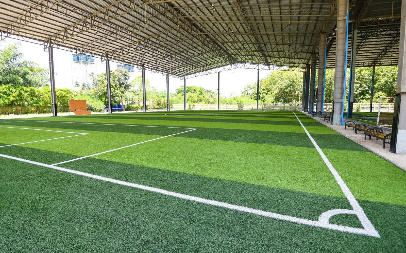 An indoor green artificial turf soccer field with white boundary lines. The area is covered by a large metal roof with supporting beams, and benches are positioned along one side. Trees and a fence are visible in the background. Licensed installers offer free consults to ensure top-quality installation.