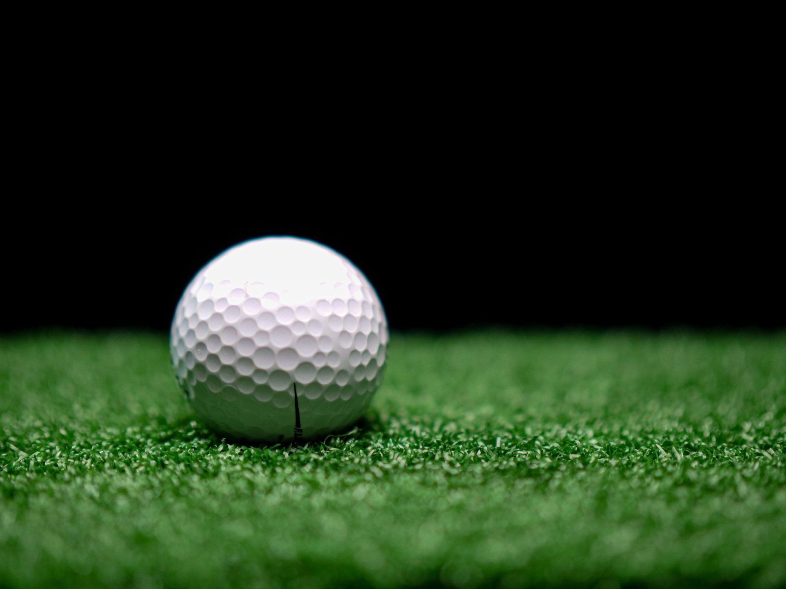 A close-up shot of a white golf ball resting on a backyard putting green in Scottsdale AZ. The background is dark, emphasizing the textured surface and dimples of the golf ball. The grass appears well-manicured, thanks to professional Golf Green Installers, highlighting the ball in the center of the image.