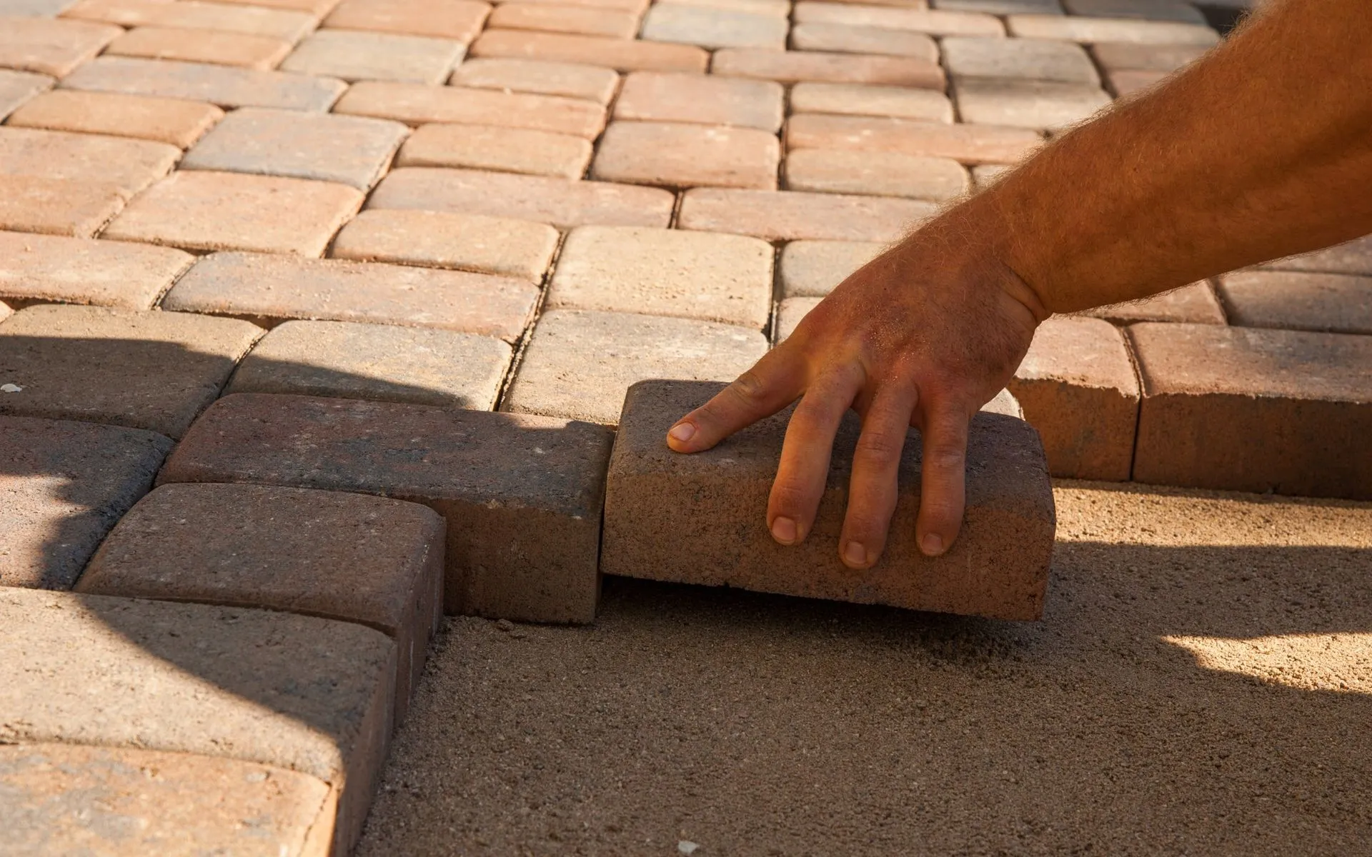 Close-up of a person's hand placing a rectangular brick onto a sand base, forming part of a neatly arranged brick pathway. The pathway, characteristic of meticulous paver installation in Scottsdale AZ, consists of uniformly aligned bricks in shades of red and brown.