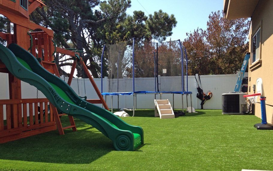 A colorful backyard playground with a green slide, wooden climbing structure, and enclosed trampoline. There is also a basketball hoop on the side, a ladder against a fence, and trees in the background. The ground is covered with pet-friendly turf by the top-rated artificial grass installer offering free estimates.