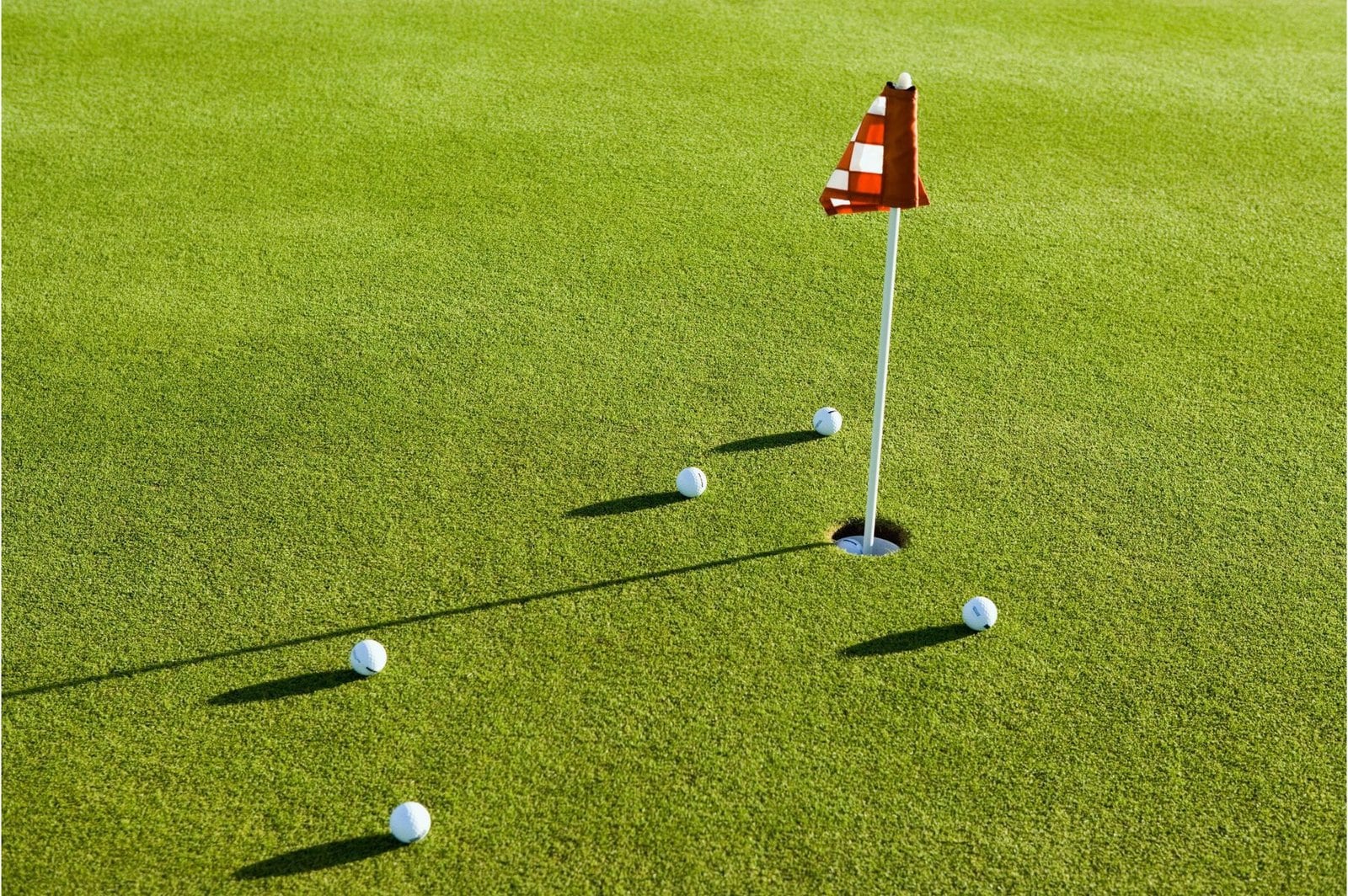 A golf hole with a flag on an immaculate green field of artificial turf. Five golf balls are positioned around the hole, casting shadows on the well-manicured grass. The flag is red and white striped.