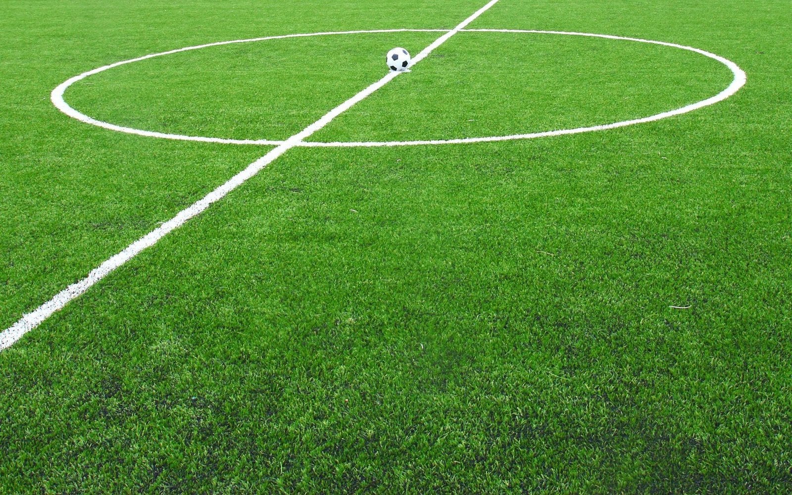 A soccer ball sits stationary at the center of a vibrant green soccer field. The white center circle and half-line are clearly marked around the ball, contrasting with the perfectly manicured artificial turf.