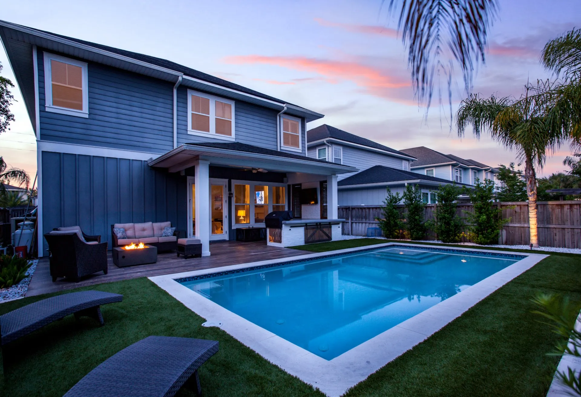 A serene backyard in Scottsdale, AZ features a two-story blue house with white trim, a covered patio with outdoor furniture, and a fire pit. The focal point is a rectangular swimming pool surrounded by lush artificial grass. Palm trees and neighboring houses are visible in the background.