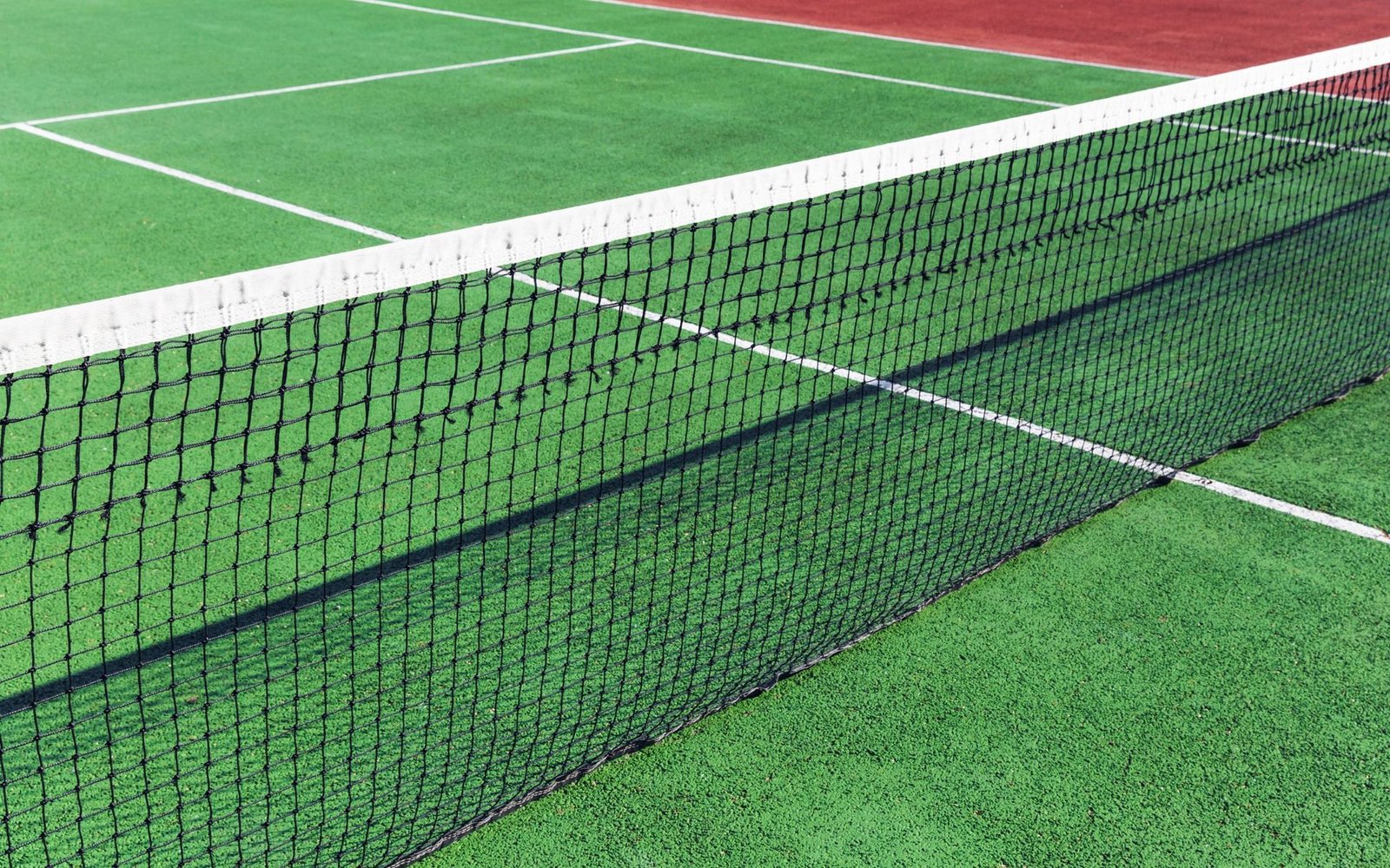 A close-up view of a tennis court featuring a black net and green artificial turf installed by licensed installers. The white court lines contrast with the green surface, and part of the court in the background shows a reddish color. The sunlight creates shadows on the net and court. Free consults available.