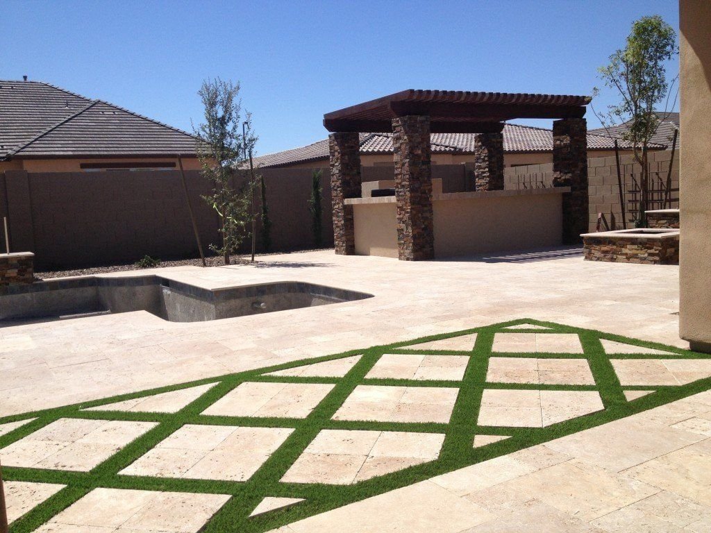 A spacious backyard in Scottsdale, AZ, with a stone patio featuring crisscrossing strips of Scottsdale Artificial Turf, an empty rectangular pool, a pergola-covered outdoor kitchen with stone pillars, and a background of beige-walled buildings and clear blue sky.