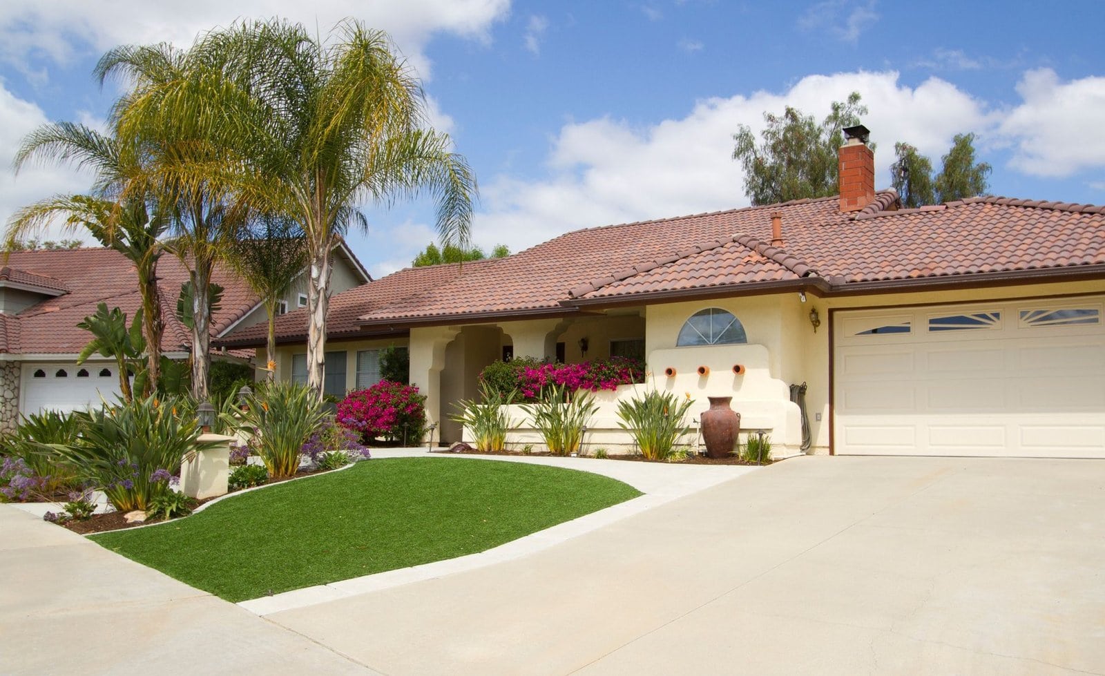 A single-story house with a tiled roof, featuring a well-maintained front yard with a small patch of artificial turf bordered by a concrete driveway. Palm trees, flowering shrubs, and a large potted plant near the entrance add charm to this home designed by licensed installers.